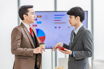 Asian male professional businessmen colleague in formal business suit take coffee break standing holding disposable paper cup and tablet computer talking have conversation together in front monitor