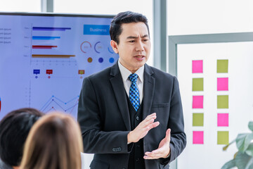 Asian professional successful businessman manager standing showing company growth profit target graph chart presentation on computer monitor to young male and female colleagues in office meeting room