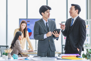 Asian male professional businessmen colleague in formal business suit take coffee break standing holding disposable paper cup and tablet computer talking have conversation together in front monitor