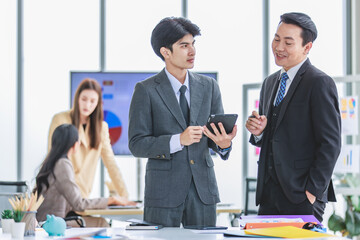 Asian male professional businessmen colleague in formal business suit take coffee break standing holding disposable paper cup and tablet computer talking have conversation together in front monitor