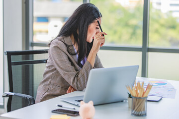 Asian young pretty thoughtful concentrated female professional successful businesswoman in formal business suit sitting holding pen at chin thinking about customer job task solution ideas with laptop