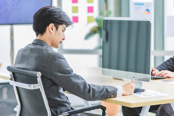 Asian young professional successful handsome male businessman in gray formal business suit sitting at working desk holding pen writing note paperwork document with computer monitor in company office