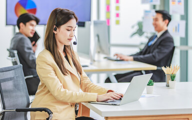 Millennial Asian young happy female customer service operator helpdesk employee staff in formal business suit sitting wearing microphone headset working talking helping customer online via laptop - obrazy, fototapety, plakaty