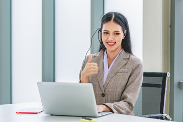 Asian young beautiful happy female businesswoman customer service call center operater in formal business suit sitting at working desk chat online with client via microphone headset in company office