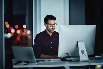 Staying late to get it done. Shot of a young businessman using a computer at his desk during a late...