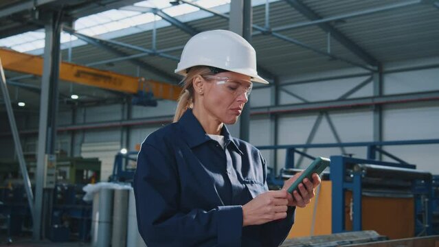At sunlight woman with helmet, uniform and goggles uses a mobile phone stand in factory. In the background details of a metalworking project