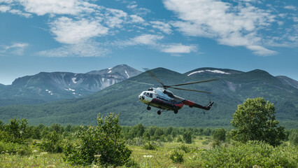 Fototapeta na wymiar The helicopter lands on a green meadow. Lush grass, wildflowers, trees all around. A picturesque mountain range against the blue sky. Kamchatka. Nalychevo