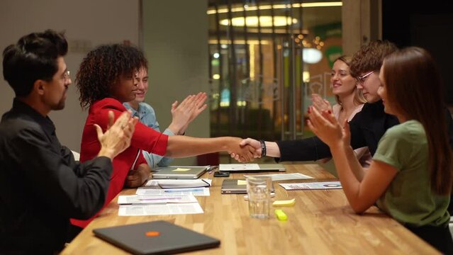 Side view of happy young African business woman and Caucasian business man shaking hands signing contract at group negotiations getting team applause after making business deal, slow motion.