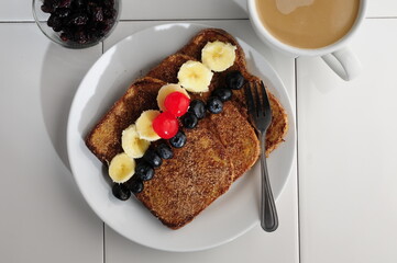 breakfast with french toast with sugar and cinnamon and slices of banana, blackberries and cherries on top, on a white plate and a cup of hot creamy coffee on the side