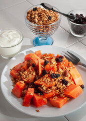 A breakfast with papaya granola and blackberries on a white plate and on a white table, with a cup of granola and blackberries on the side, vertical side view