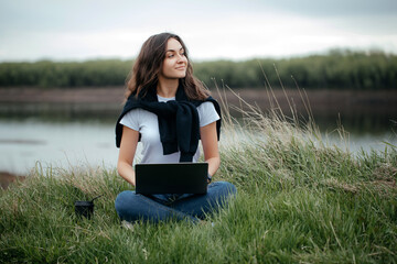 beautiful freelance girl works remotely outside the city sitting on a track with a laptop near the river enjoying the fresh air. concept of distance learning, online learning.