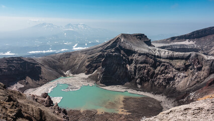 Naklejka premium Amazing turquoise acid lake on top of an active volcano. Melted snow at the water's edge. The layered structure of the steep slopes of the crater is visible. Blue sky. Kamchatka. Gorely Volcano