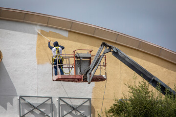 Man painting a yellow wall white using a paint sprayer while standiong on a manlift - obrazy, fototapety, plakaty