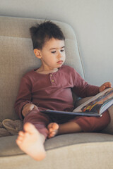 Little boy reading a text book while sitting comfortable on sofa at home. Family, childhood, leisure and people concept.