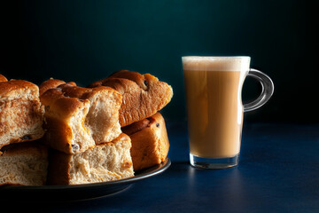 A glass cup of hot coffee Moka cappuccino with a black plate with cinnamon raisin bread or rolls on a dark blue table with a dark Turquoise background horizontal side view.