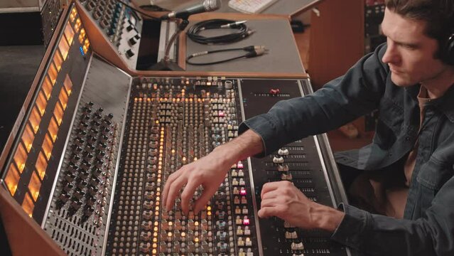 Tilt up slowmo of serious young male music producer in headphone using mixing console while adjusting sound before record at professional music studio