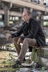 A young short-haired man in brown jeans and a denim shirt is smoking sitting in some abandoned hangar. Probably a laborer sat down to rest