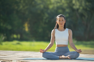 Naklejka na ściany i meble Woman on a yoga mat to relax in the park at mountain lake. Calm woman with closed eyes practicing yoga, sitting in Padmasana pose on mat, Lotus exercise, attractive sporty girl in sportswear.