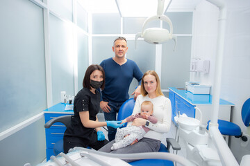 Obraz premium Young parents and their daughter visit a dentist to examine milk teeth. Baby at the first appointment with the dentist. Inspection of the formation of jaws, eruption of milk teeth.