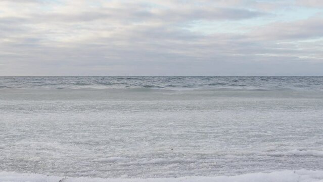 Static shot of ice covered sea waves rolling in on beach. Cloudy blue sky and white palette