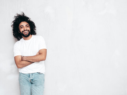 Handsome smiling hipster  model.Sexy unshaven Arabian man dressed in summer stylish white t-shirt clothes. Fashion male with long curly hairstyle posing near grey wall in studio. Isolated