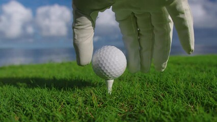 Male hand in a golf glove puts the ball on the field on the background of the blue ocean. Close up