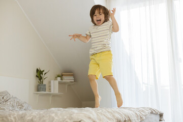 Fototapeta na wymiar A little boy joyfully jumps on the bed in the bedroom. Fun games at home. Children's activity and games with parents