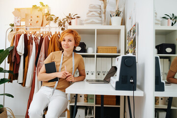 Short-haired seamstress posing next toher workdesk with a notebook