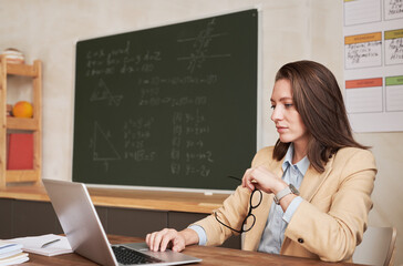 Side view portrait of young female teacher sitting at desk in school classroom and using laptop,...