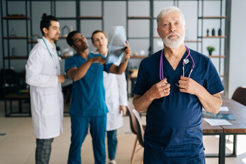 Portrait of confident senior aged surgeon in blue uniform standing in medical meeting office, looking at camera. Multiethnic team of physician working in background, discussing MRI scan of patient.