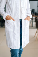Close-up vertical cropped shot of unrecognizable female doctor in white uniform holding hands in pockets standing in hospital office. Practitioner woman posing at workplace. Concept of medical work.