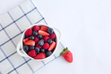 A white bowl of berries on a plaid napkin on the table. Summer snack. Light food photography on a white background top view