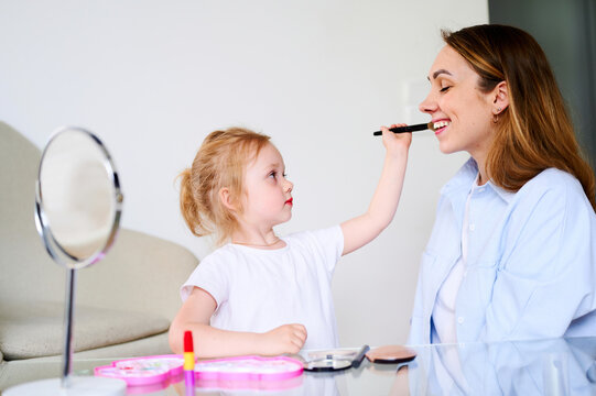 Mom and daughter play laugh and paint their lips with red lipstick. Mother teaches the child to do makeup. The relationship between parents and children