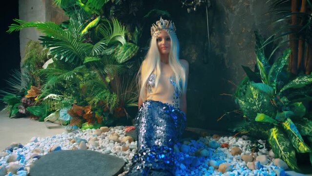 fantasy woman queen mermaid sits on floor in studio. Rain shower, green plant modern bathroom interior. Silver fish tail creative design costume fin, blonde nymph goddess girl princess, crown on head