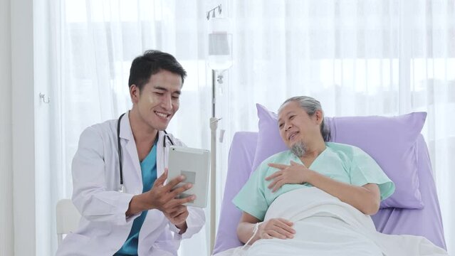 Asian male doctor explains symptoms to elderly male patient using tablet in the ward at the hospital