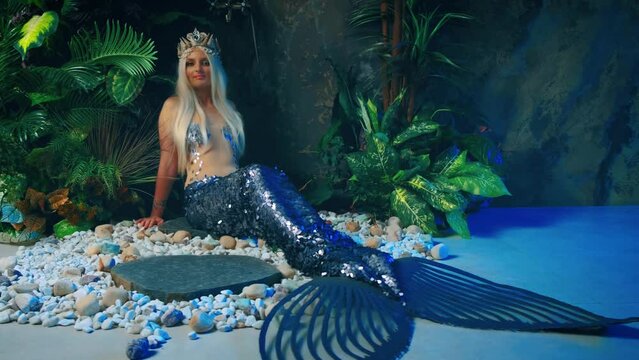 fantasy woman queen mermaid sits on floor in studio. Rain shower, green plant modern bathroom interior. Silver fish tail creative design costume fin, blonde nymph goddess girl princess, crown on head
