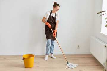 Full length portrait of attractive slim woman wearing white t shirt and apron posing with cleaning equipment and washing floor in her living room, doing household chores.