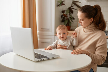 Portrait of dark haired adorable woman wearing beige jumper posing in kitchen with her infant daughter and working on laptop, looking after baby during maternity leave, working online.