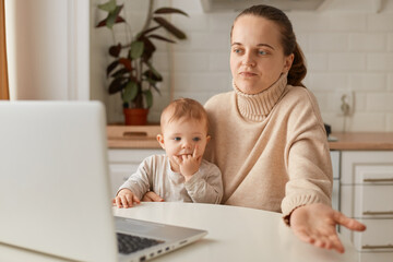 Fototapeta na wymiar Image of puzzled dark haired woman with ponytail wearing beige jumper posing in kitchen and having online conversation, holding toddler daughter in hands, shrugging, being not sure.