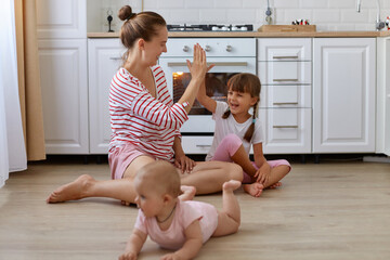 Indoor shot of smiling woman wearing striped casual style shirt sitting on floor in kitchen with her children, cute little baby crawling, mother giving five to her daughter.