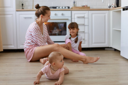 Image of happy family, woman wearing striped casual style shirt sitting on floor in kitchen with daughters, toddler baby crawling, mother talking with elder kid.