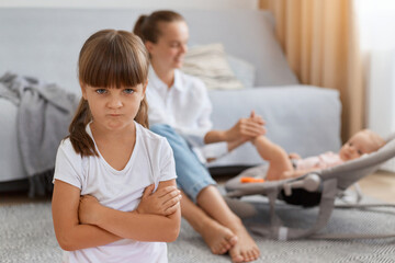 Portrait of offended sad little girl standing with folded hands, looking at camera with sadness, jealous mother to baby sister, woman in white shirt and jeans with kid in rocking chair on background.