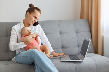 Horizontal shot of woman with bun hairstyle wearing white shirt and jeans sitting on cough, holding baby daughter in hands, talking via mobile phone and typing on laptop.