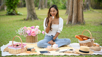Beautiful Asian young woman sipping a tea, making a picnic in the green park.