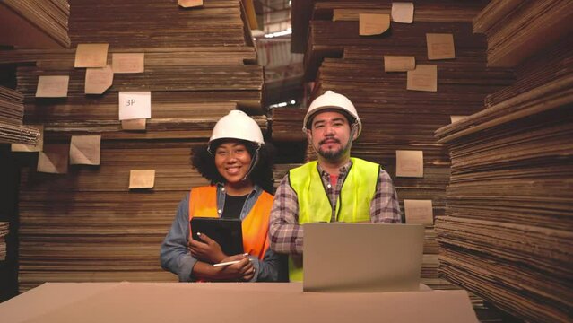 Engineer asian and african woman wearing safety helmet and vest holding clipboard and take note on the paper in the automotive part warehouse. Factory for the manufacture and processing of paper.