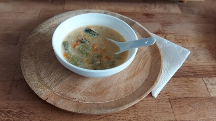 Gemüsesuppe. Suppenschüssel und Löffel aus weißem Porzellan. Weiße Serviette am Boden der Suppentasse. Holzsockel. Suppe serviert auf einem Holzteller. Vegetarisches Gericht. Karotten und Kohlrabi.