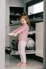 Fototapeta na wymiar Cute toddler girl wearing casual clothes, holding clean plates. Kid helping in the kitchen.