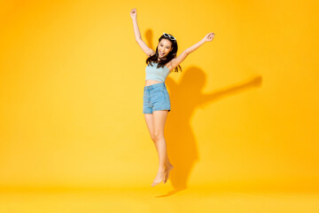 Fototapeta na wymiar Happy energetic Asian woman wearing summer outfit jumping and raising hands in studio isolated yellow color background