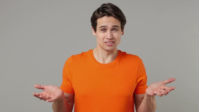 Fun confused shy shamed young brunet man 20s years old wears orange t-shirt looking camera spreading hands say oops ouch oh omg i am so sorry isolated on plain grey wall background studio portrait