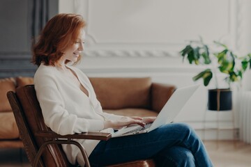 Smiling woman freelancer sits in armchair, keyboards on laptop computer, uploads necessary material...